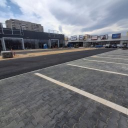 Firma Brukarska Roboty Ziemne Dariusz Karykkowski - Ukończony parking z ciemnoszarej kostki brukowej o falistym wzorze, z białymi liniami, przed nowymi lokalami handlowymi i blokiem mieszkalnym.
