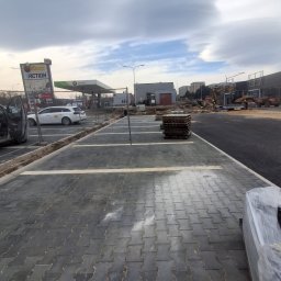 Firma Brukarska Roboty Ziemne Dariusz Karykkowski - Parking z ciemnoszarej kostki brukowej w jodełkę, z białymi pasami, obok stos materiału. W tle stacja Amic, szyldy Biedronki/Action i budowa z koparką.