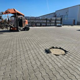 Firma Brukarska Roboty Ziemne Dariusz Karykkowski - Duży plac wyłożony szarą kostką brukową typu H, z pomarańczową układarką do kostki i częściowo odsłoniętą studzienką. W tle magazyn i palety z materiałem.