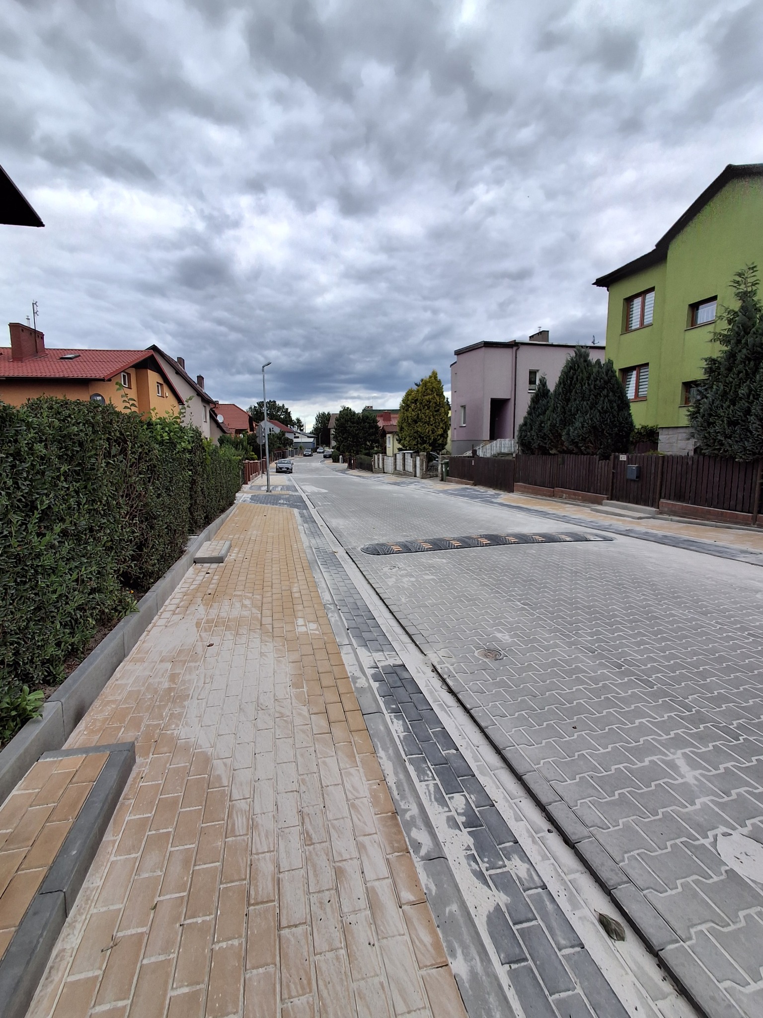 Ulica z nową kostką brukową w odcieniach szarości i beżu, z progiem zwalniającym. Po lewej chodnik z kostki, po prawej domy mieszkalne. Pochmurne niebo.