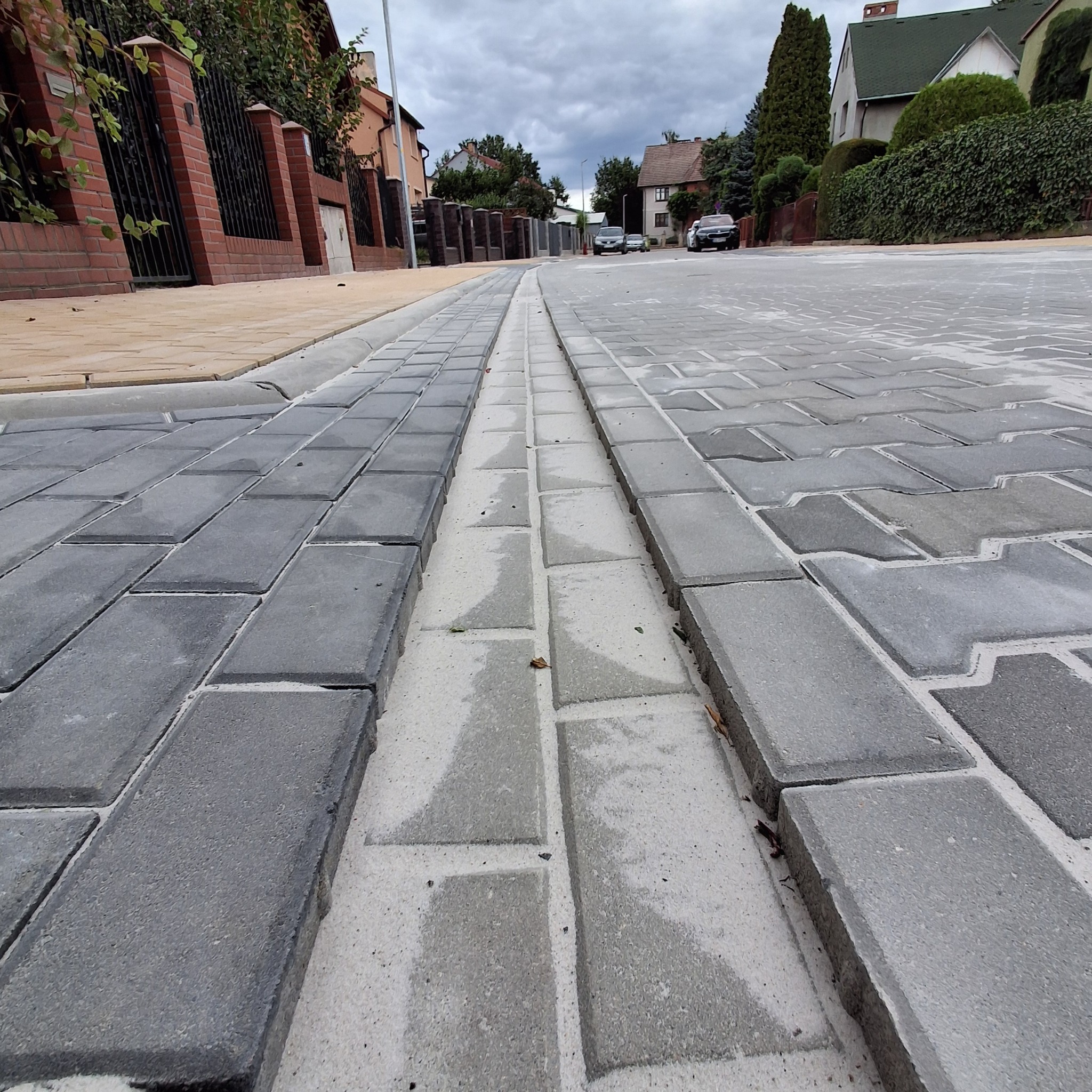 Ułożona kostka brukowa w szarych odcieniach, z centralnym odpływem liniowym, na tle zabudowy jednorodzinnej i pochmurnego nieba. Widok z perspektywy chodnika.