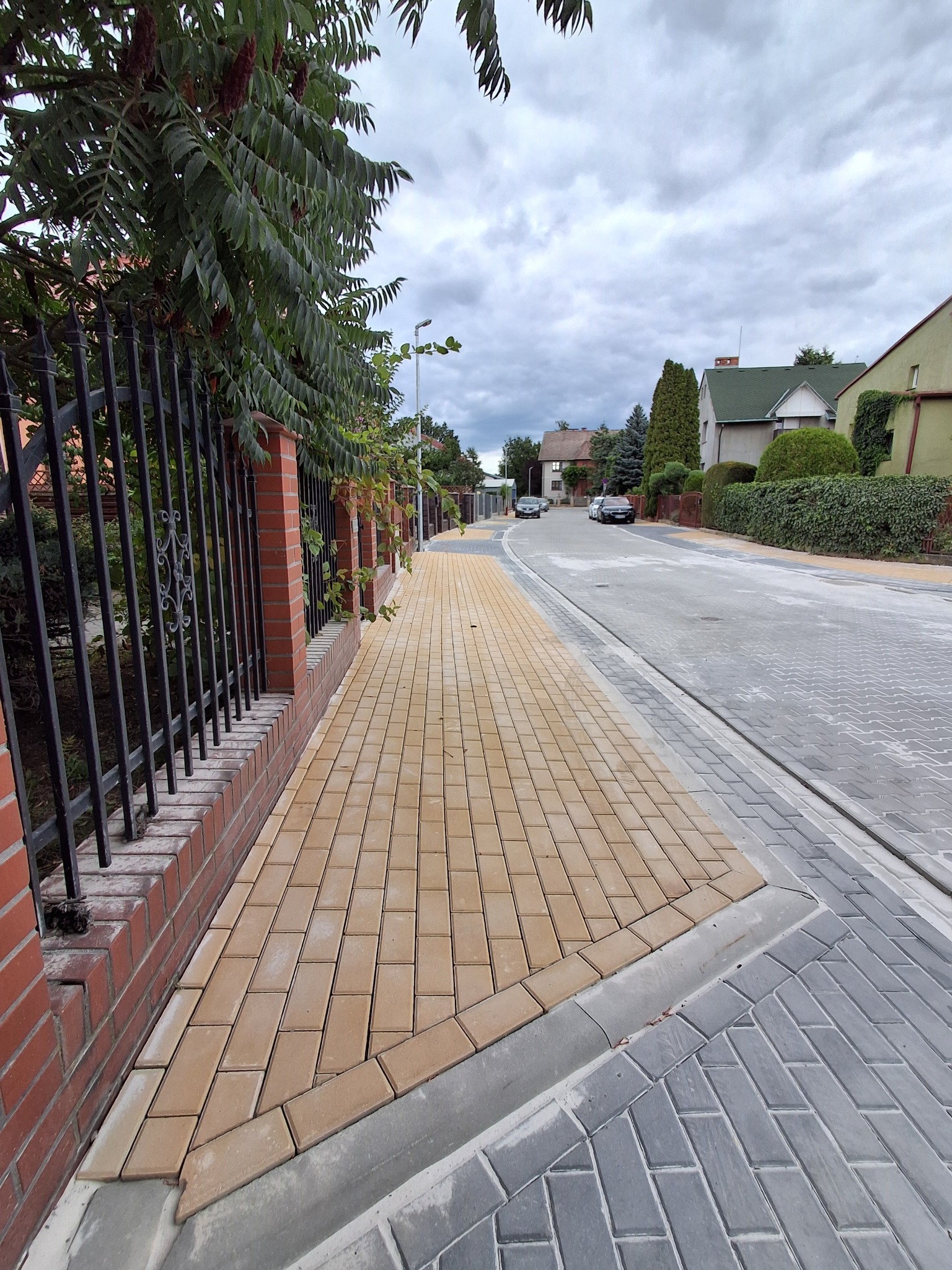 Żółta kostka brukowa ułożona wzdłuż ogrodzenia z cegły i metalu, przechodząca w szarą kostkę na ulicy. Widok na domy i pochmurne niebo.