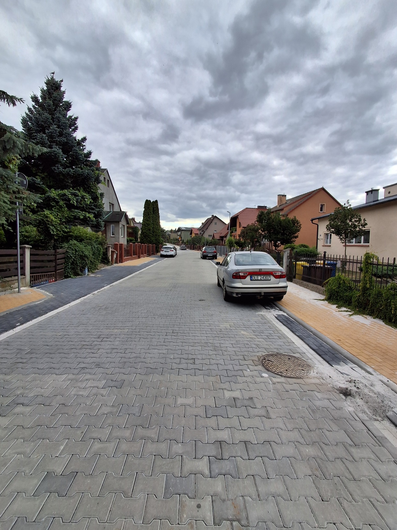 Ulica osiedlowa z szarej kostki brukowej, obrzeża z żółtej kostki. W tle domy jednorodzinne i pochmurne niebo. Zaparkowany samochód po prawej stronie.