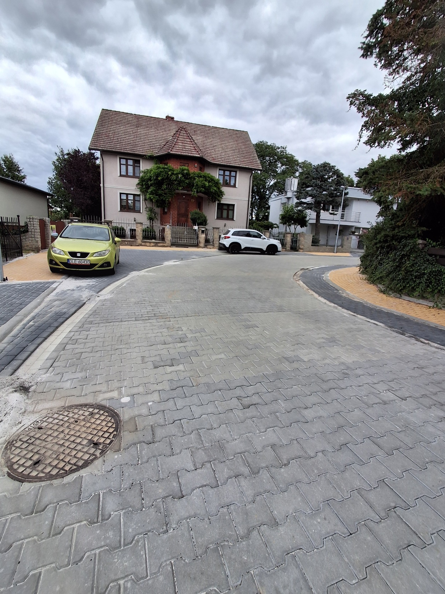 Nowa, szara kostka brukowa na drodze osiedlowej z widocznymi domami i samochodami. Żółte obrzeża chodnika. Studzienka kanalizacyjna wkomponowana w nawierzchnię.