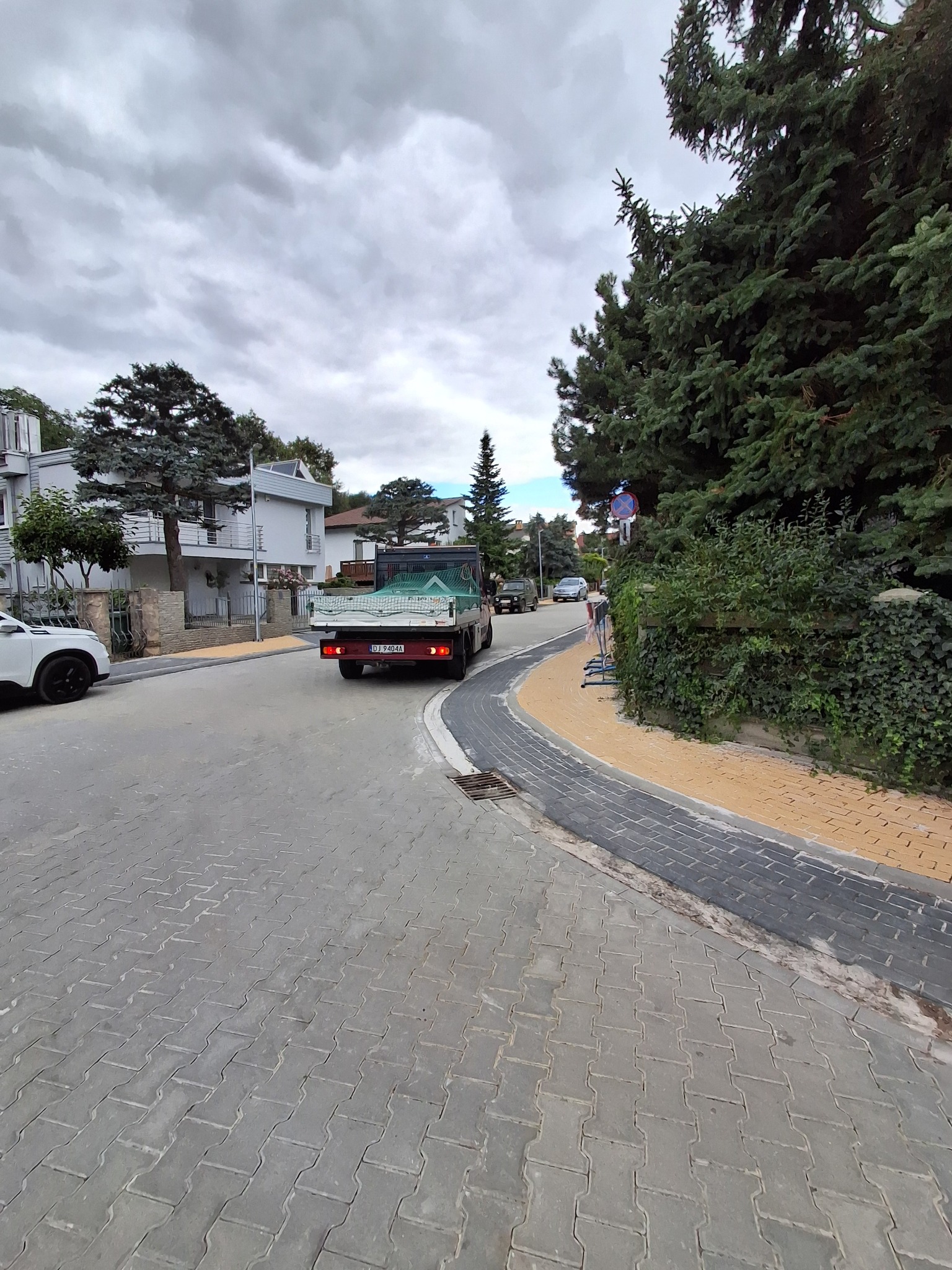 Ulica z ułożoną szarą kostką brukową w jodełkę, obok żółty pas piasku. W tle zaparkowany biały samochód i jadąca ciężarówka z zieloną siatką na pace.