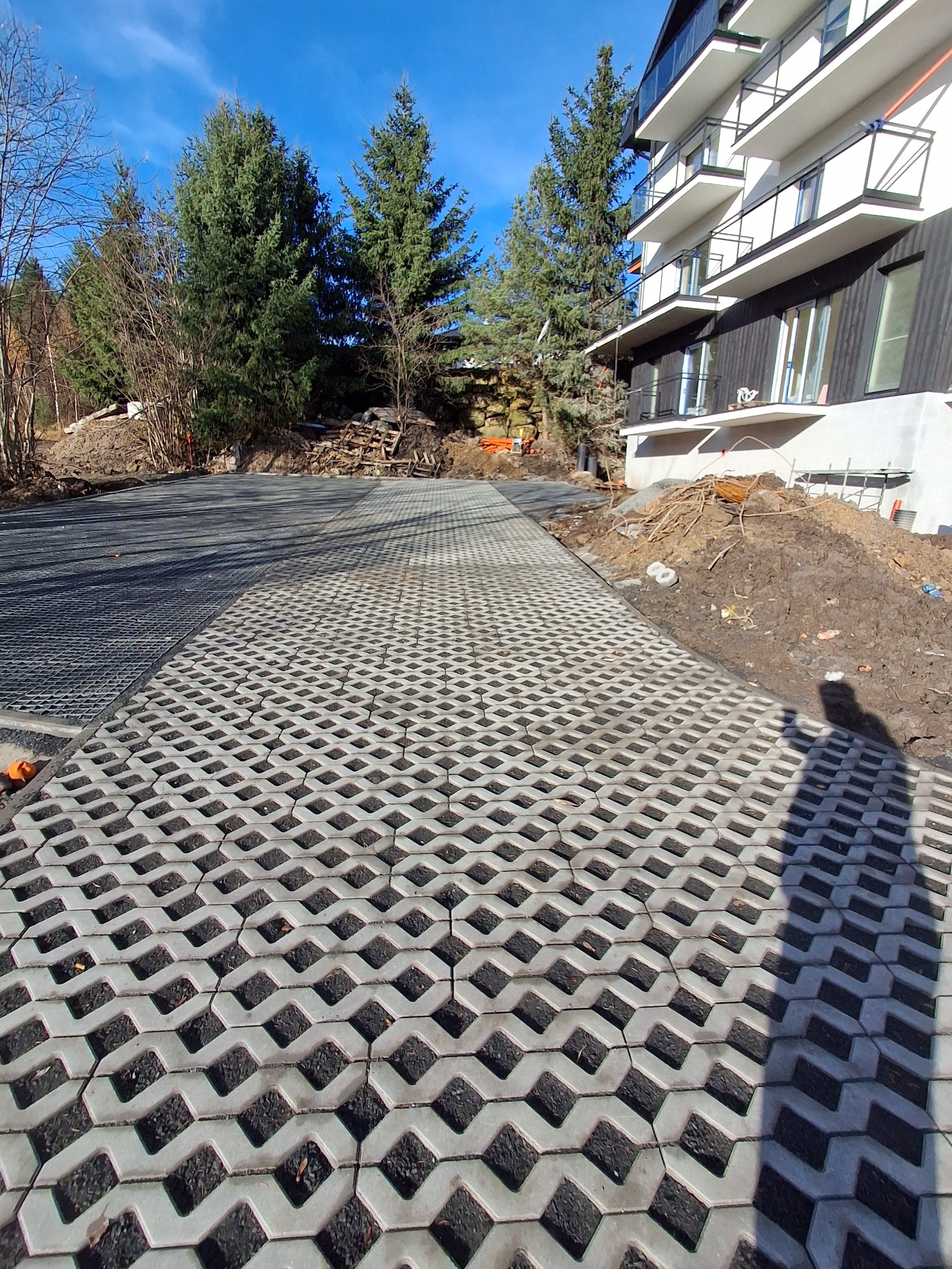 Ułożona szara ażurowa kostka na podjeździe przed budynkiem z balkonami, widoczne drzewa i fragment cienia osoby. Słoneczny dzień, błękitne niebo.