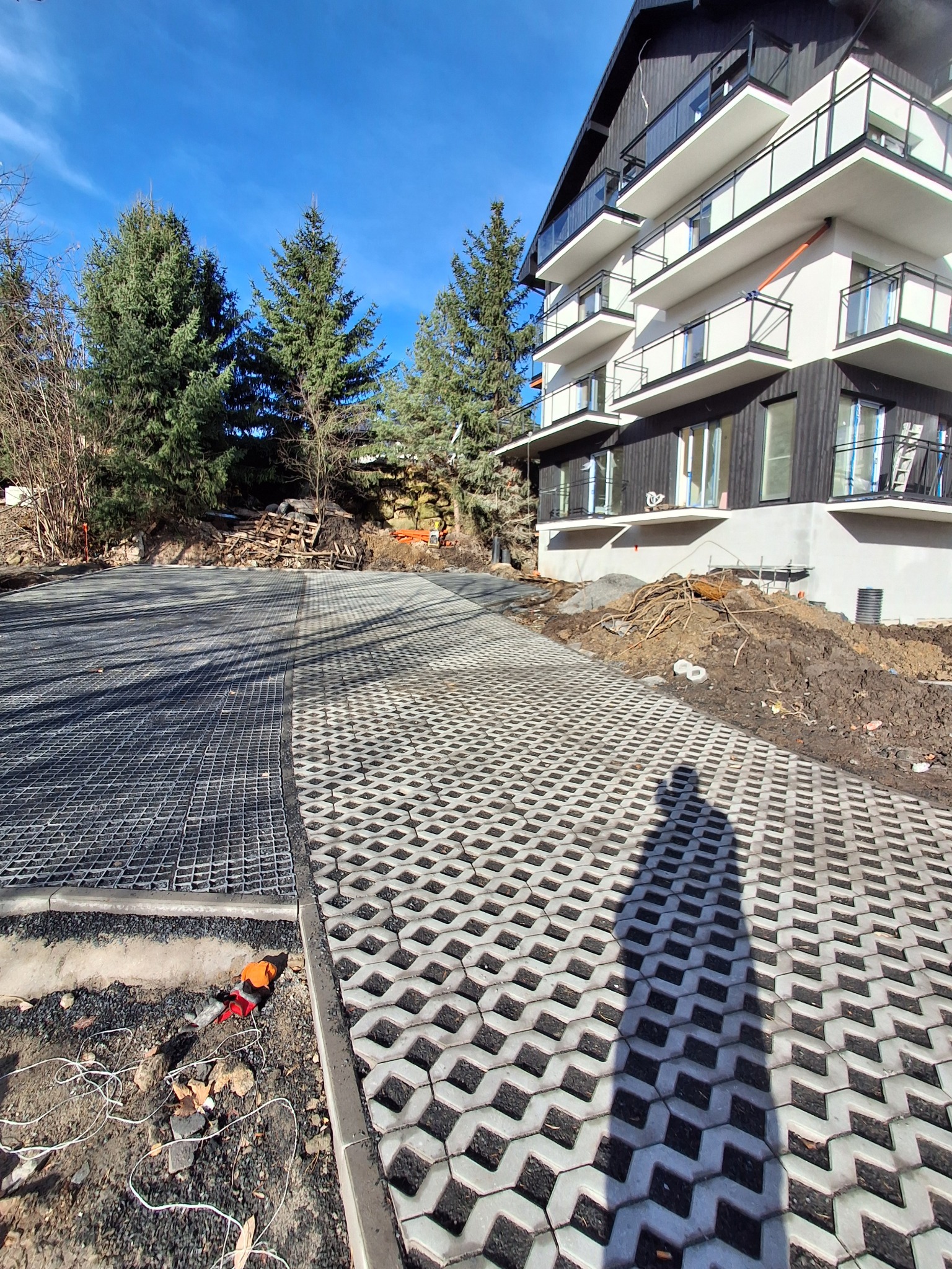 Układanie przepuszczalnej kostki brukowej na podjeździe nowego apartamentowca. Część powierzchni wyłożona, część przygotowana pod ułożenie. Widoczne narzędzia.