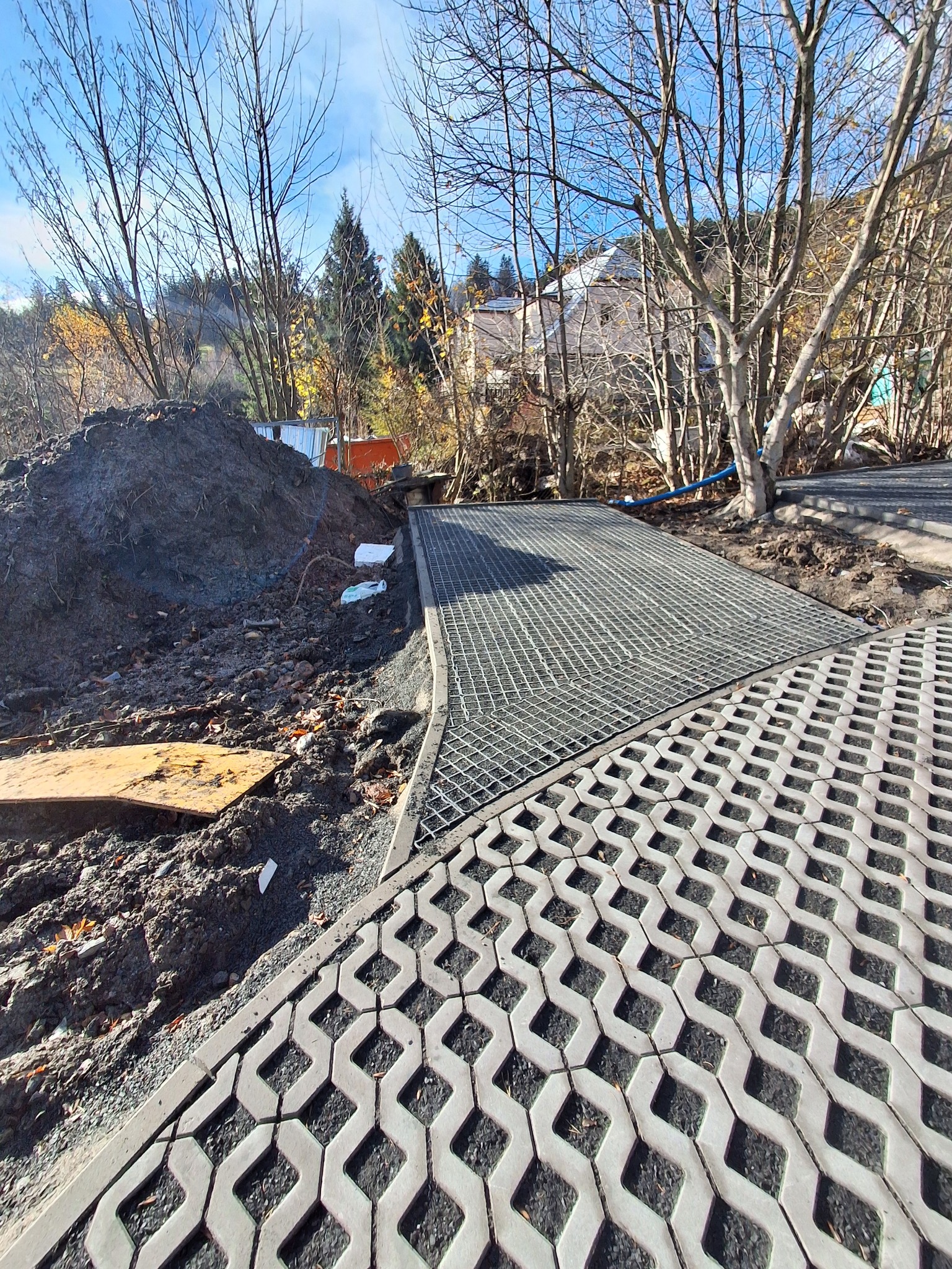 Fragment terenu z ułożoną ażurową kostką brukową i metalową kratą, obok pryzma ziemi. Widok na wzgórze z drzewami i zabudowaniami w tle, słoneczny dzień.
