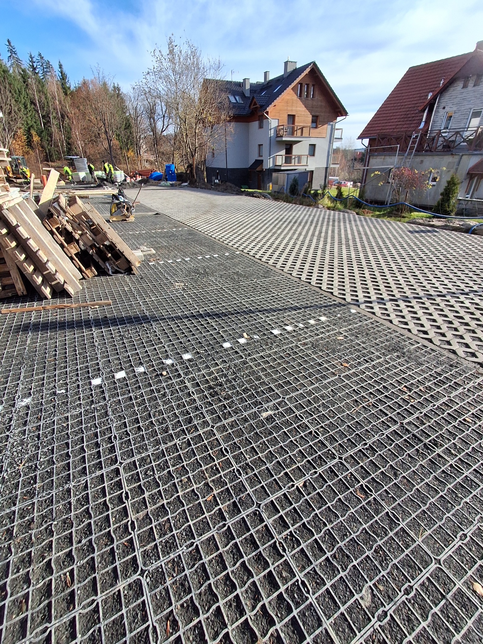 Montaż ażurowych płyt drogowych na podbudowie z kruszywa. W tle ekipa budowlana i budynki mieszkalne. Widok na częściowo wykonaną nawierzchnię.