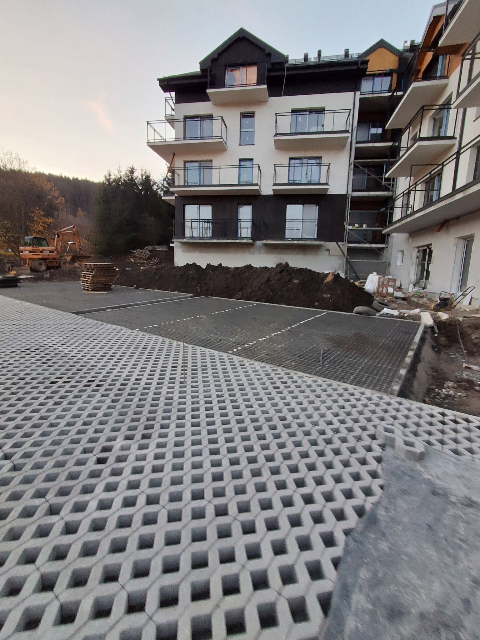 Ułożona szara ażurowa kostka na parkingu przed budynkiem mieszkalnym. W tle widoczna koparka i palety. Częściowo przygotowany teren pod dalsze prace.