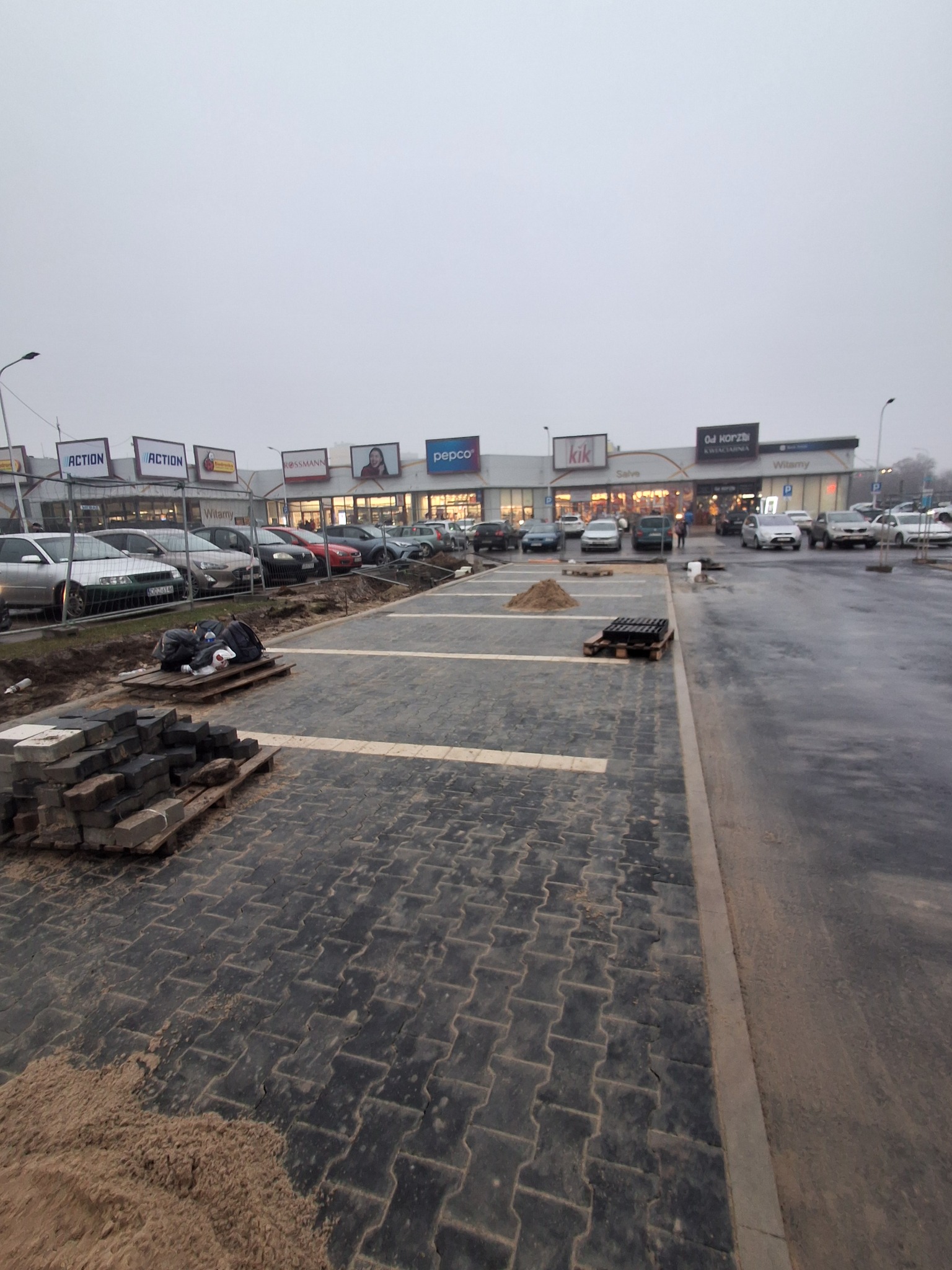 Częściowo ułożona, ciemna kostka brukowa na parkingu przed centrum handlowym w pochmurny dzień. Widoczne palety z kostką i piasek.