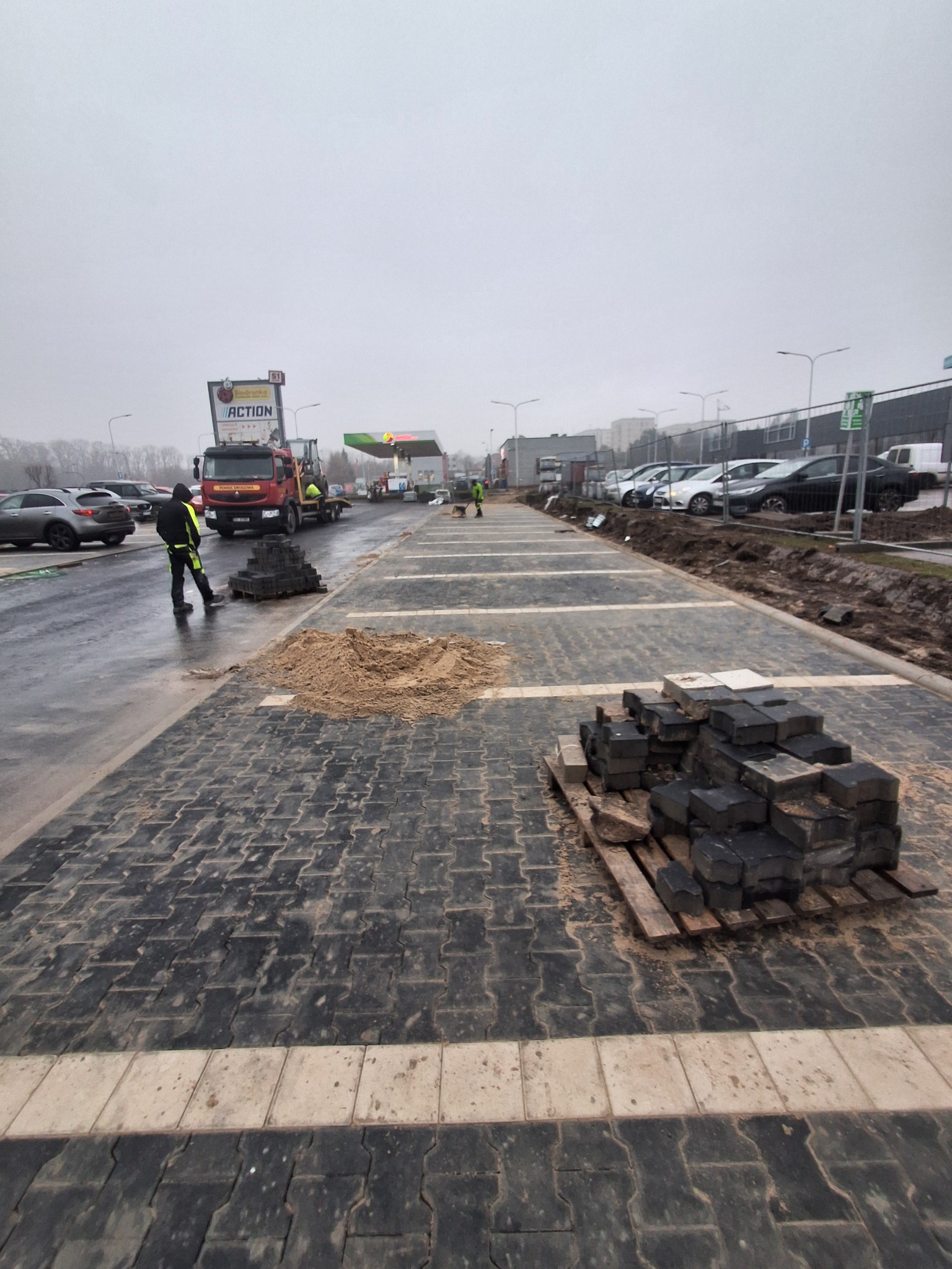 Częściowo ułożona, czarno-szara kostka brukowa z jasnymi pasami, paleta z kostką i hałda piasku. W tle pracownik w odblaskowej kamizelce i ciężarówka na parkingu.