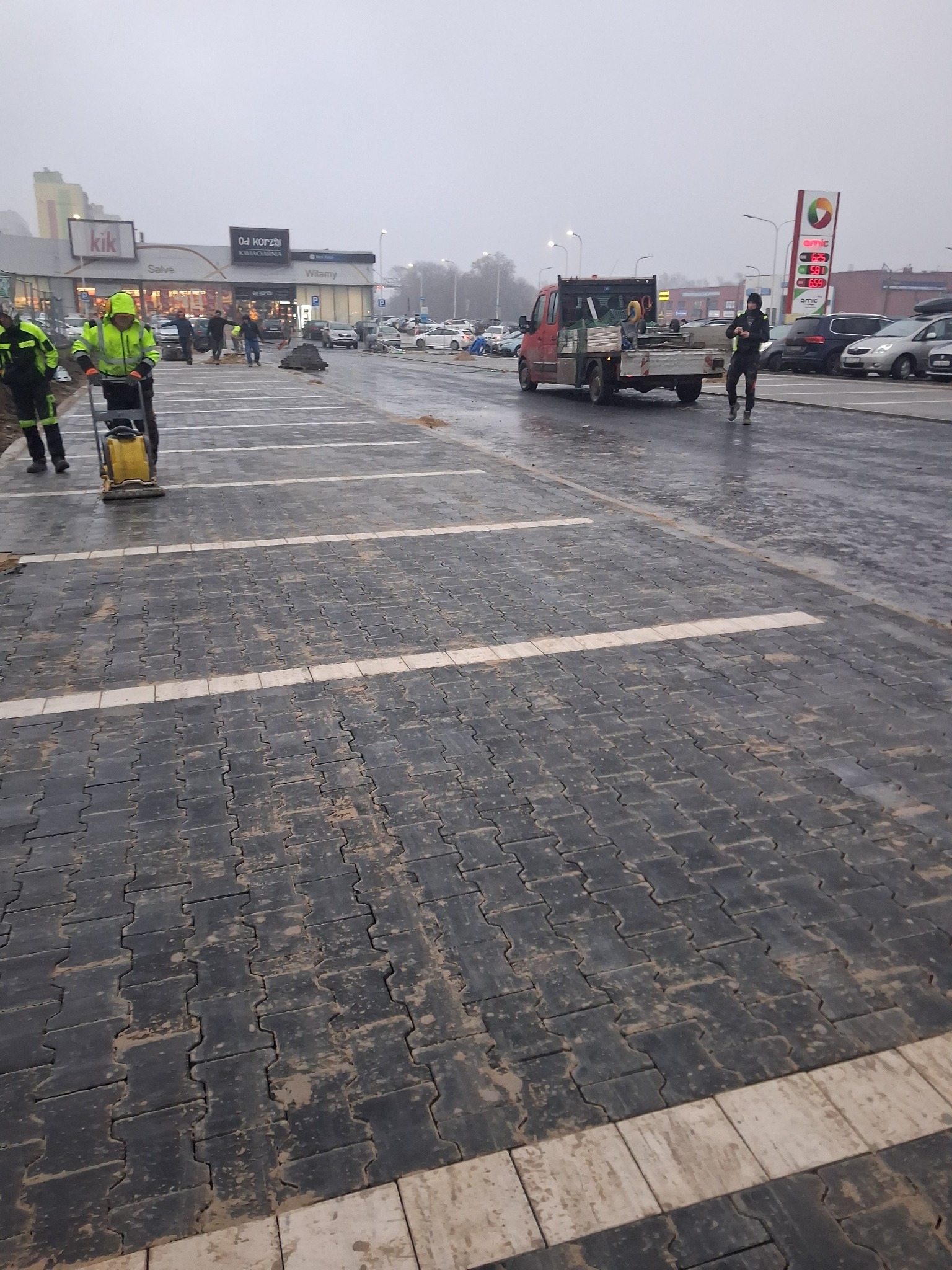 Ciemna kostka brukowa ułożona na parkingu sklepu, pracownicy w odblaskowych ubraniach, zagęszczarka w użyciu, w tle zaparkowane auta i budynki w pochmurny dzień.
