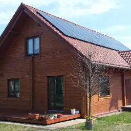 Drewniany dom z brązowymi panelami słonecznymi na dachu, tarasem i kwiatami w doniczkach, widok w słoneczny dzień.