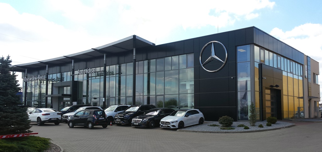 Nowoczesny budynek salonu samochodowego Mercedes-Benz z przeszkloną fasadą i dużym logo na elewacji, zaparkowane samochody przed budynkiem.