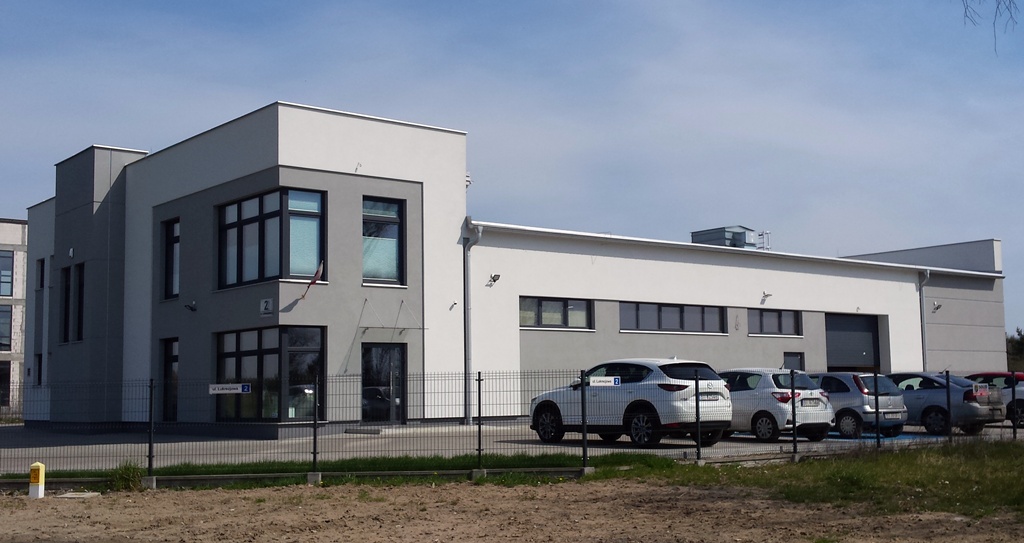 Nowoczesny budynek biurowy z szarą i białą elewacją, widoczny parking z samochodami, słoneczny dzień.