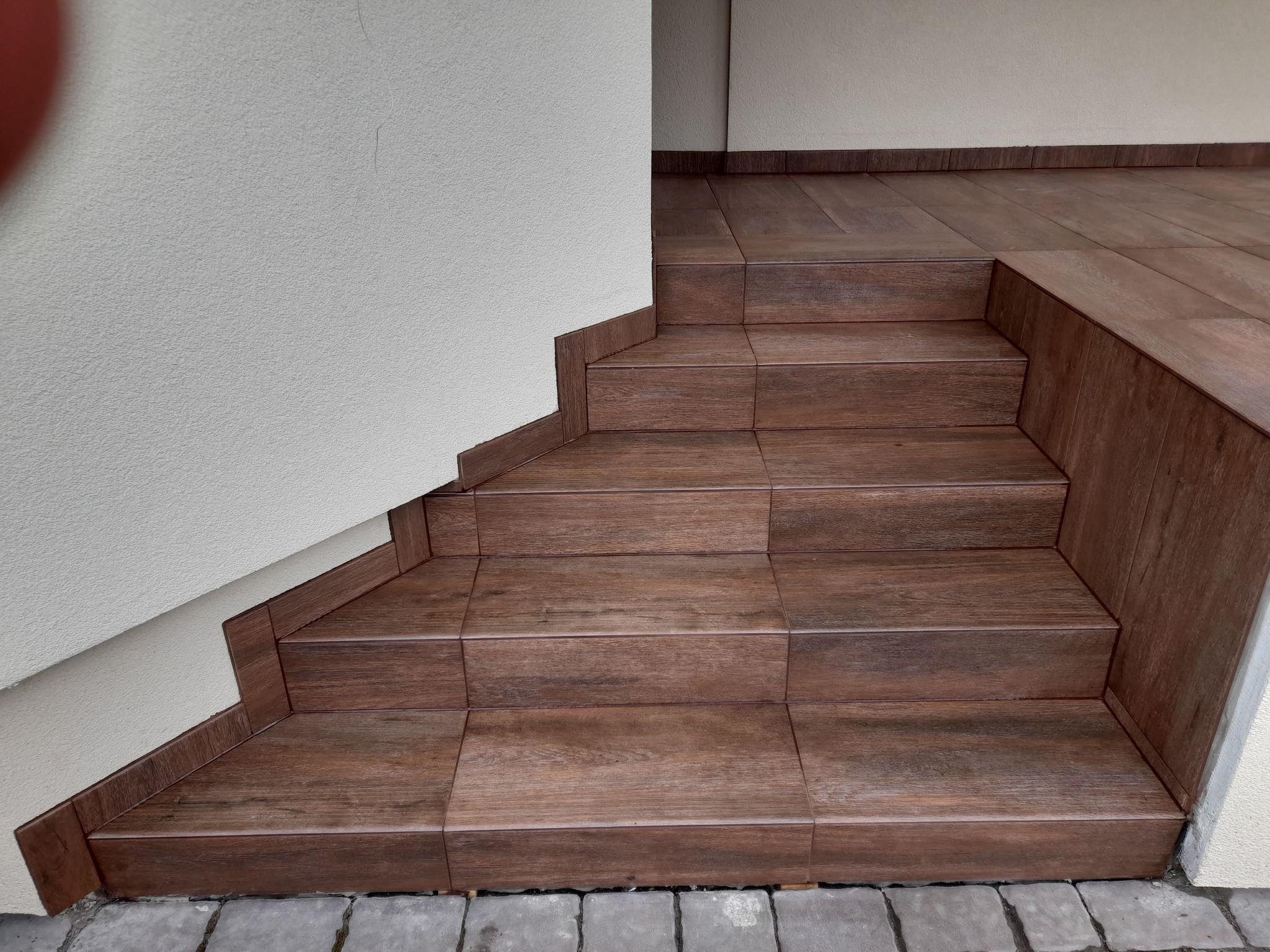 Wykończone brązowymi płytkami schody zewnętrzne z ciemną fugą, przylegające do jasnej ściany. Widoczne detale wykończenia krawędzi i dolna część muru z kostki brukowej.