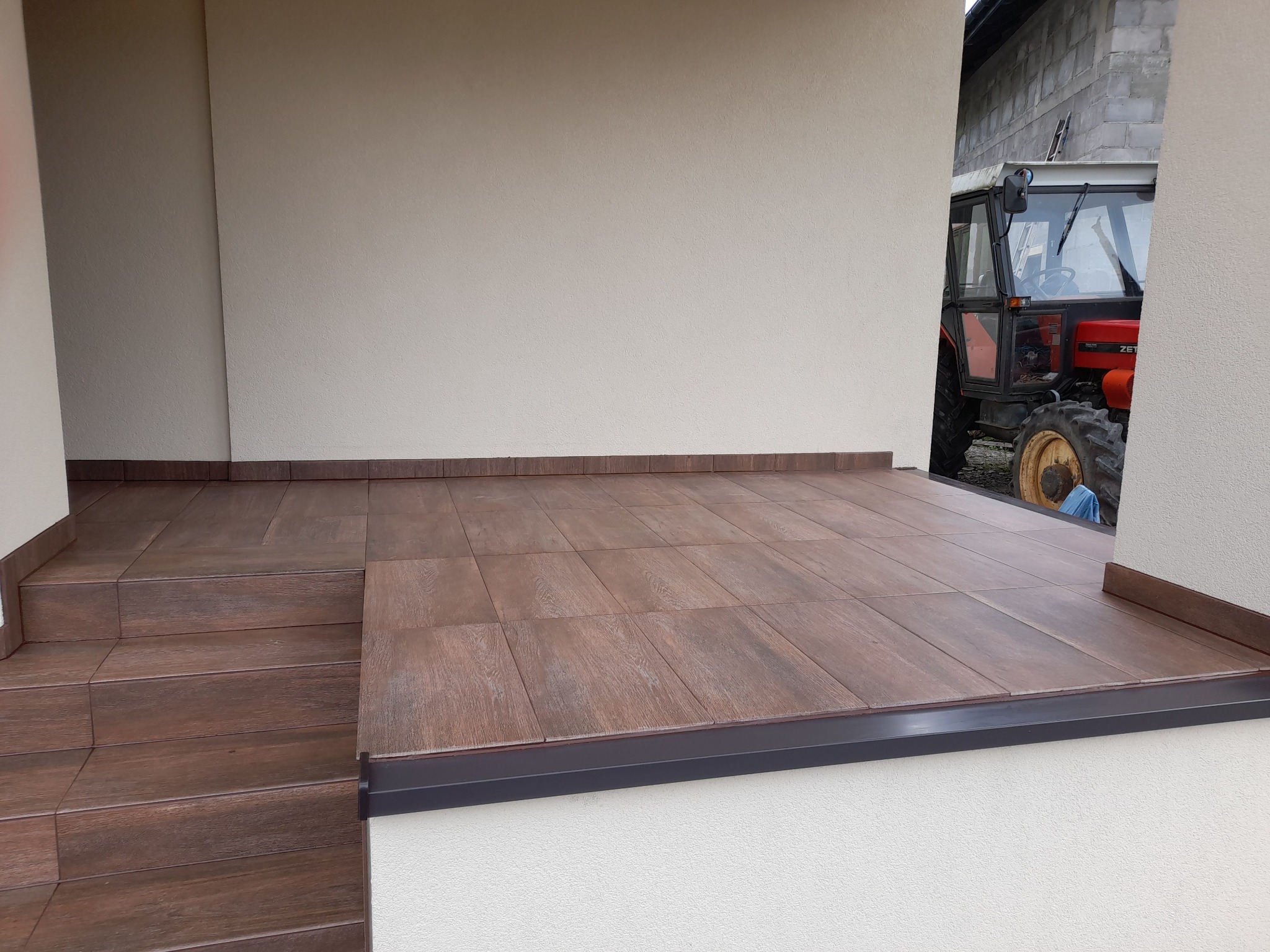 Wykończony taras z brązowych płytek imitujących drewno, z widocznymi schodami i czarnym obramowaniem. W tle fragment traktora, ściana z pustaków.