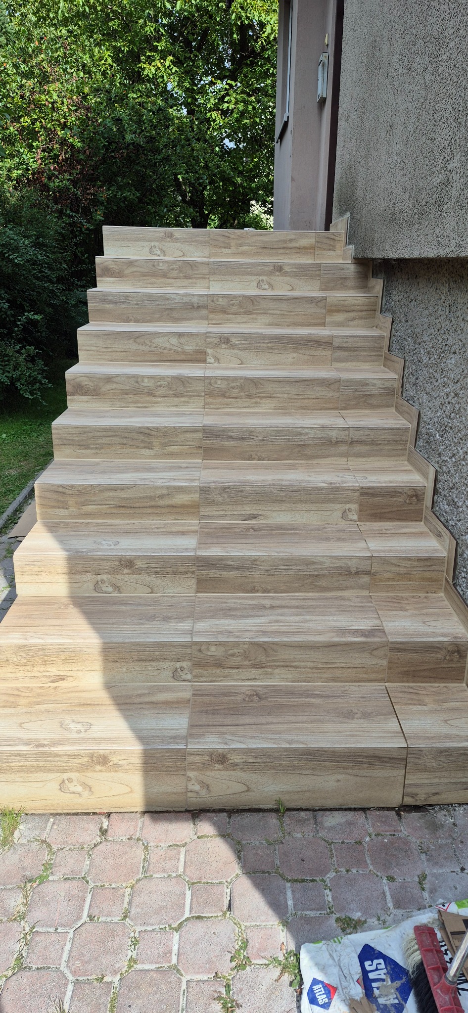 Wykończone schody zewnętrzne z płytek imitujących drewno, prowadzące do budynku z elewacją. Widoczne opakowania po materiałach budowlanych.