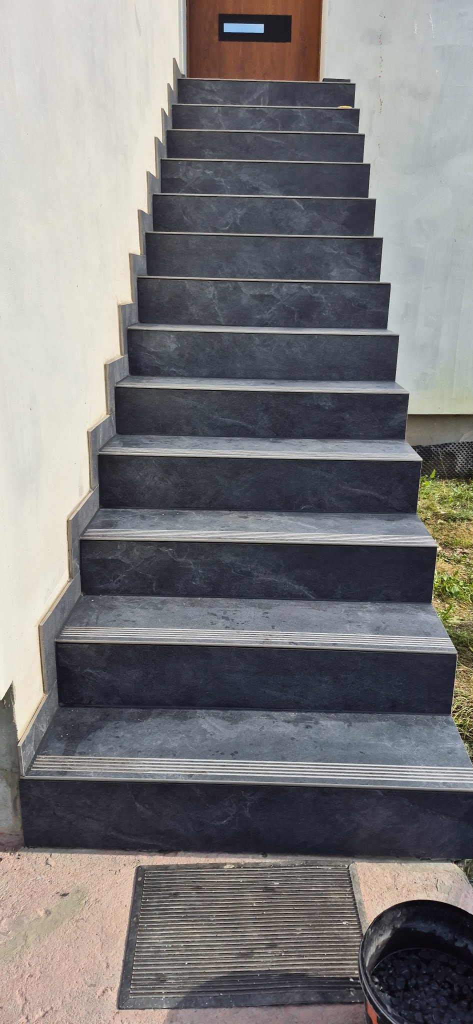 Granitowe schody zewnętrzne prowadzące do drzwi, z widoczną wycieraczką i wiaderkiem z czarnymi kamykami u dołu, oświetlone naturalnym światłem.