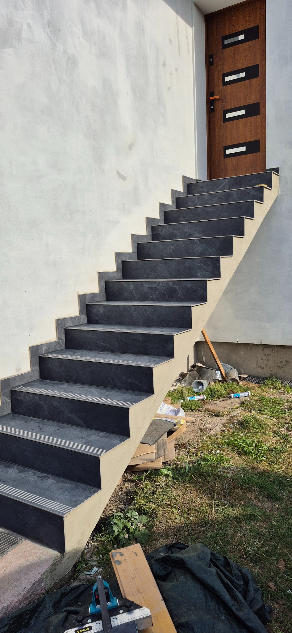 Zewnętrzne schody z ciemnymi płytkami imitującymi kamień, wykończone jasną fugą, prowadzą do brązowych drzwi wejściowych. Widoczne narzędzia glazurnicze.