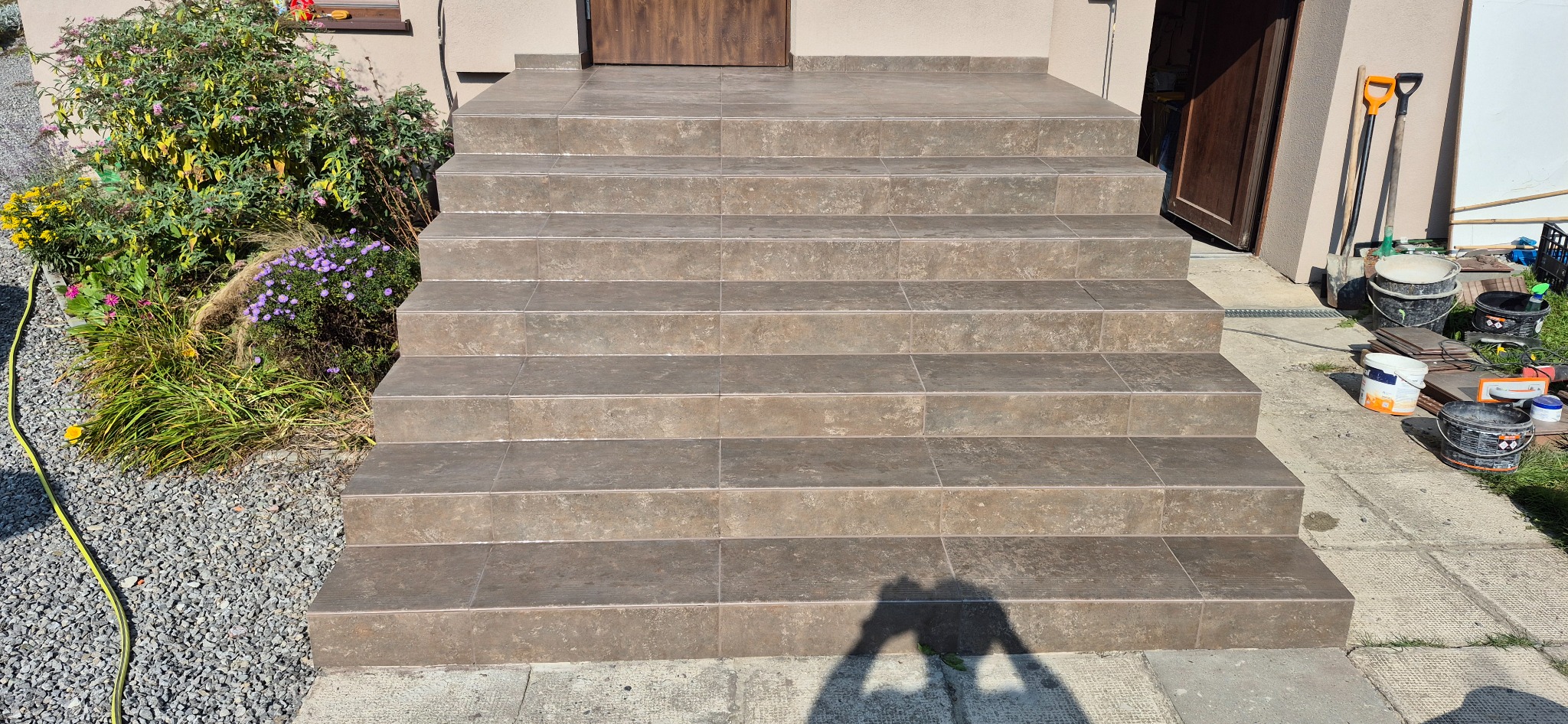 Zewnętrzne schody wejściowe wyłożone brązowymi płytkami, z widocznymi narzędziami i materiałami budowlanymi obok, oraz fragmentem zieleni po lewej stronie.