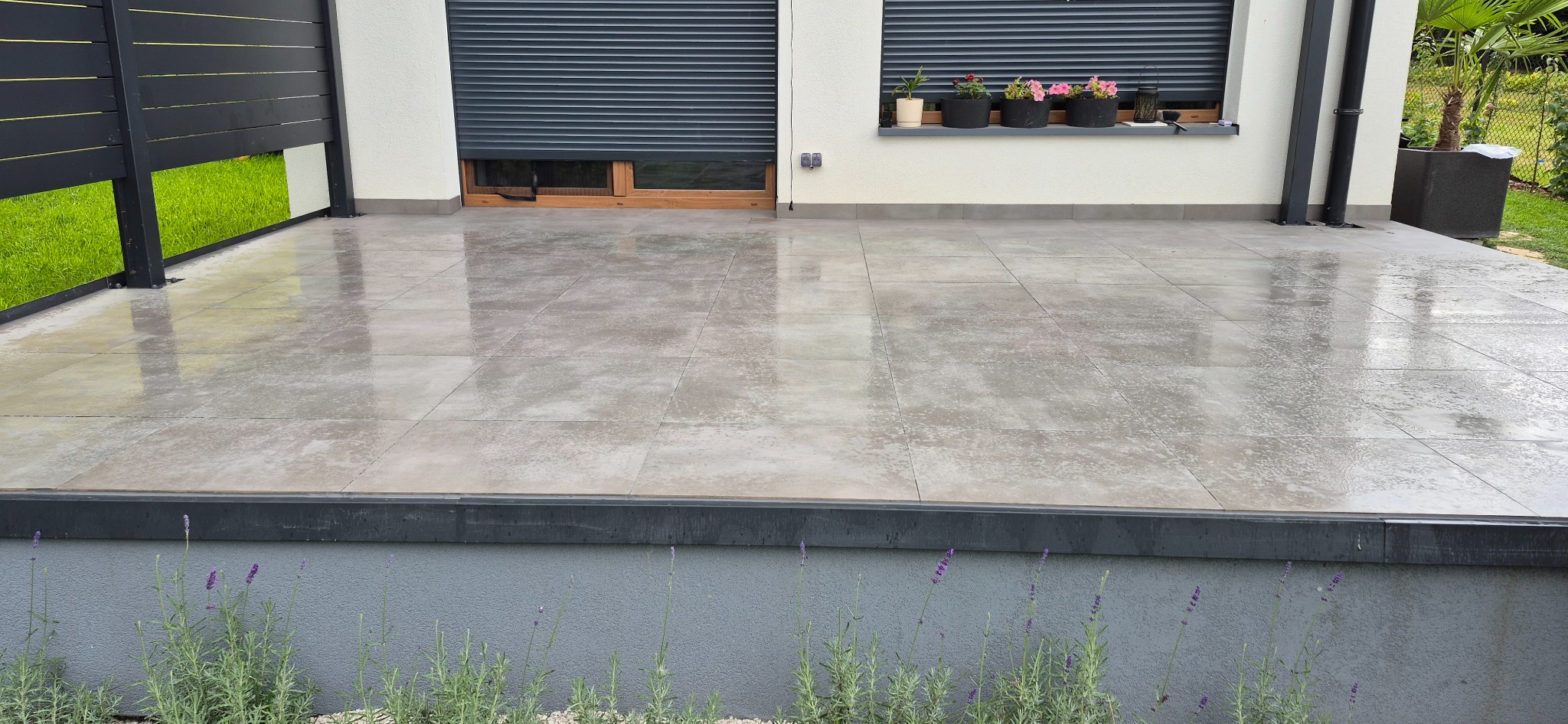 Jasnoszara, błyszcząca posadzka z płytek na tarasie, z widocznymi kwiatami w doniczkach na parapecie i fragmentem metalowego ogrodzenia. Minimalistyczny design.