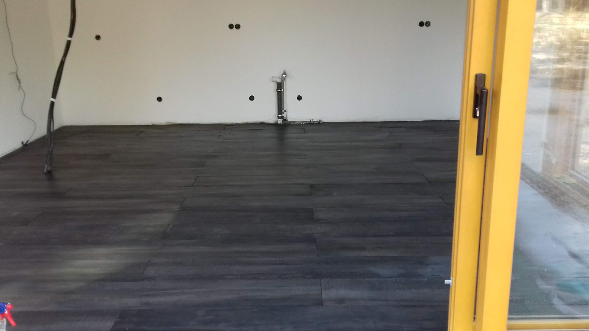 Ułożone ciemne panele podłogowe w pustym pomieszczeniu z żółtymi drzwiami balkonowymi i instalacją na ścianie. Widoczne kable i otwory w ścianie.