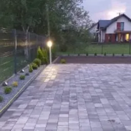 Szara kostka brukowa ułożona na posesji prywatnej, oświetlona lampami ogrodowymi wzdłuż metalowego ogrodzenia, z widokiem na dom w tle.