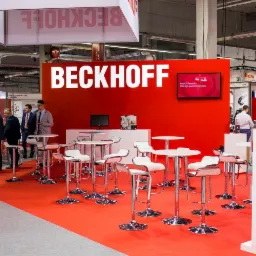 Stoisko firmy Beckhoff na targach, z czerwonym tłem i logo, stoliki barowe z białymi krzesłami, liczne osoby w garniturach.