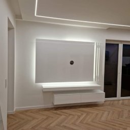 MP-ELEKTRO Marcin Palica - Nowoczesna biała ściana telewizyjna z podświetleniem LED, minimalistyczny design, parkiet w jodełkę na podłodze, widoczne okno balkonowe.