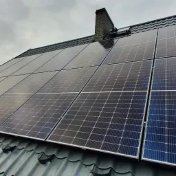 Panele fotowoltaiczne na dachu z blachy trapezowej, widoczny komin i słup energetyczny w tle.