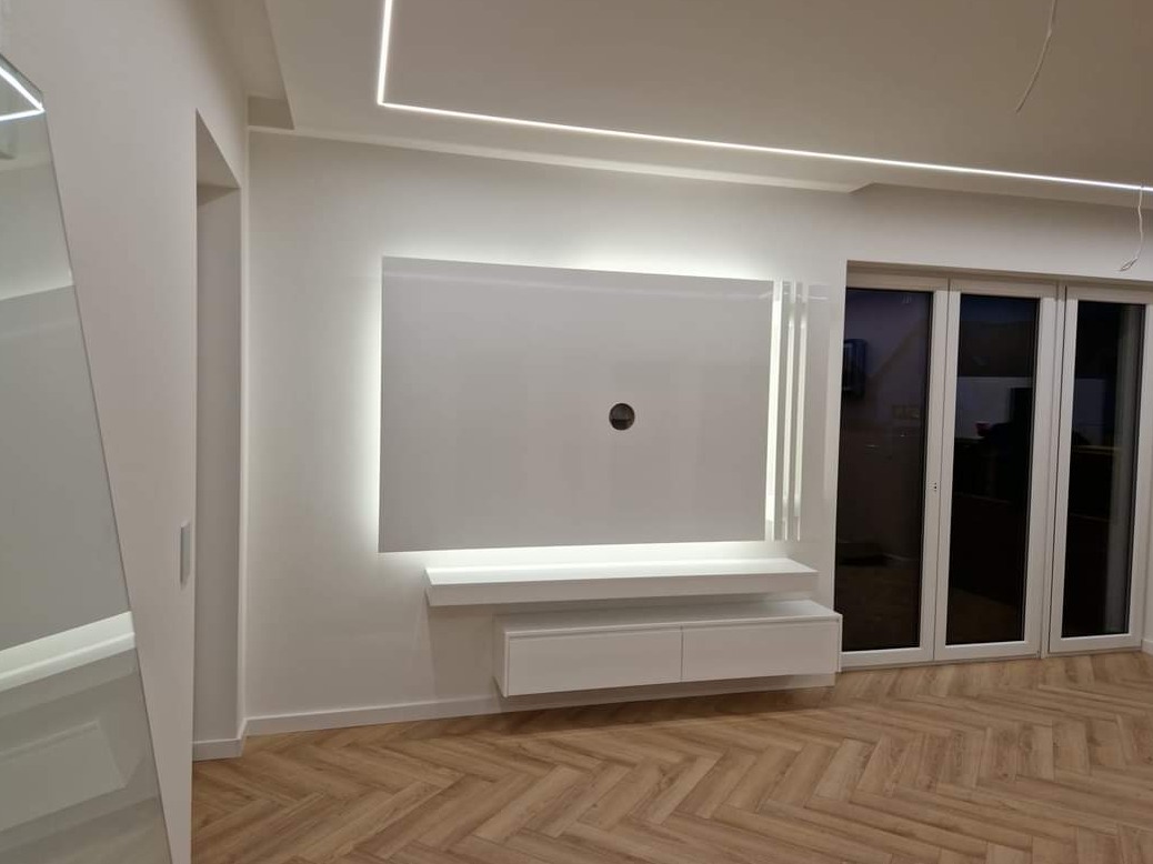 Nowoczesna biała ściana telewizyjna z podświetleniem LED, minimalistyczny design, parkiet w jodełkę na podłodze, widoczne okno balkonowe.