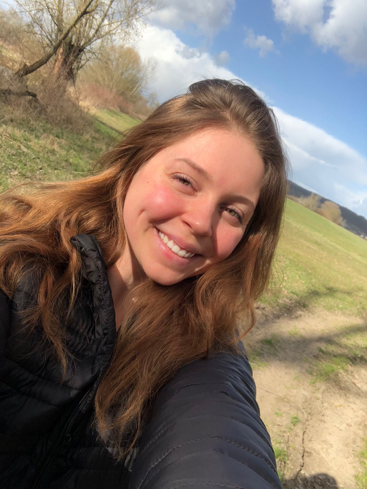 Portret uśmiechniętej młodej kobiety w czarnej kurtce na tle zielonego pola i błękitnego nieba z chmurami.