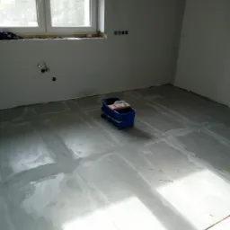 Pok&oacute;j w trakcie prac przygotowawczych do wykończenia, z widocznymi śladami po szpachlowaniu na podłodze i ścianach, oraz niebieskim wiaderkiem z narzędziami.