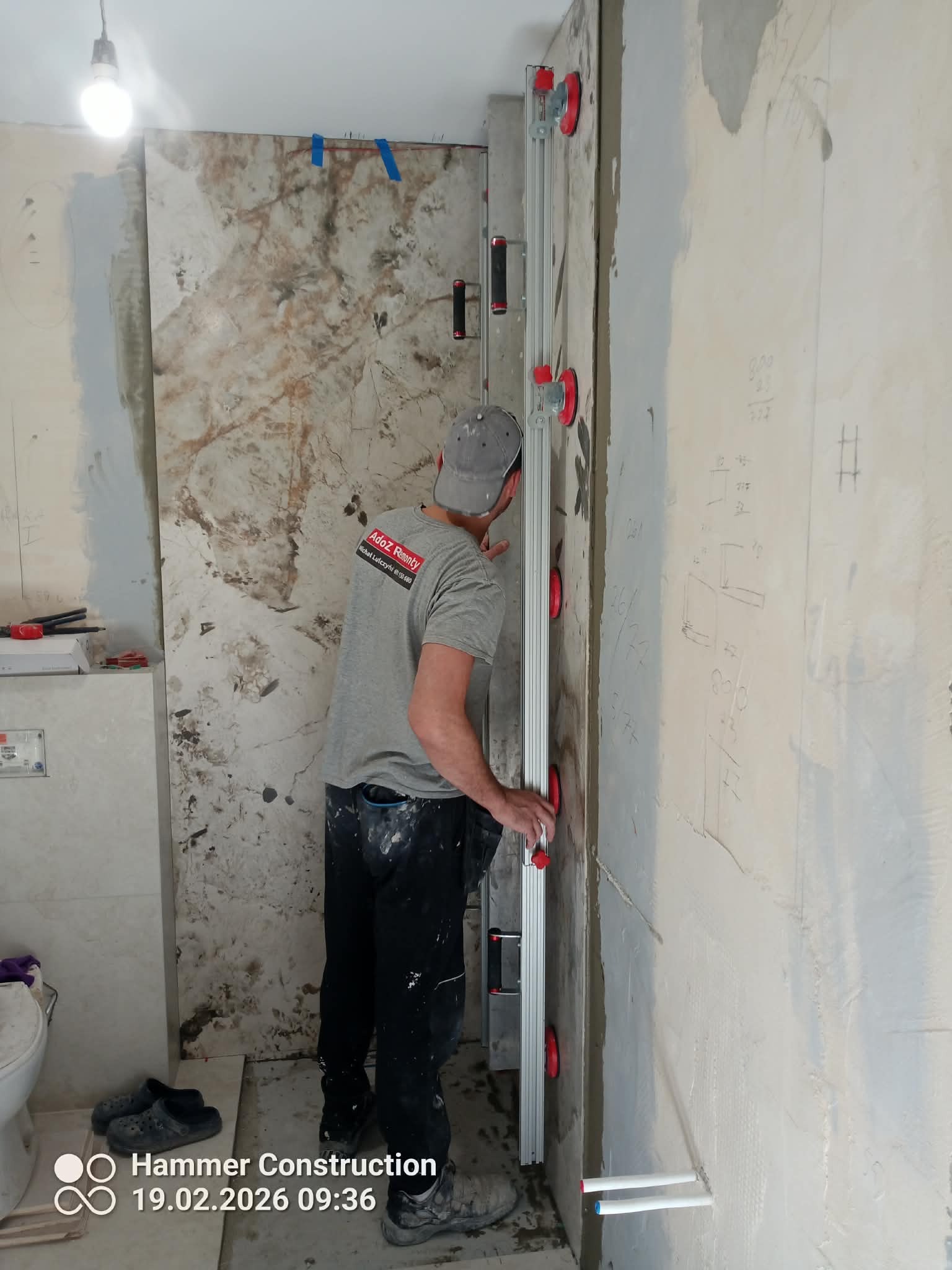 Glazurnik układa wielkoformatową płytę w łazience, używając specjalistycznego narzędzia z przyssawkami. Widoczne przygotowane ściany i instalacje.