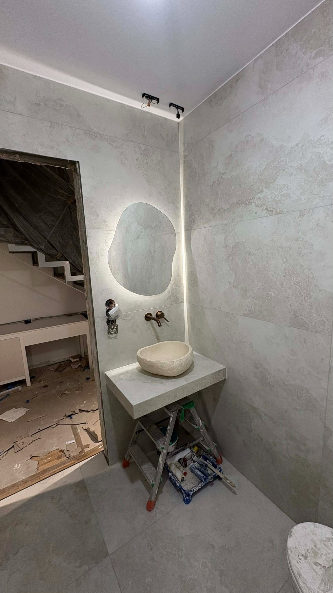 Remont łazienki: Umywalka w stylu kamienia na blacie z szarych płytek, podświetlane lustro o nieregularnym kształcie. W tle widoczna drabina i narzędzia malarskie.