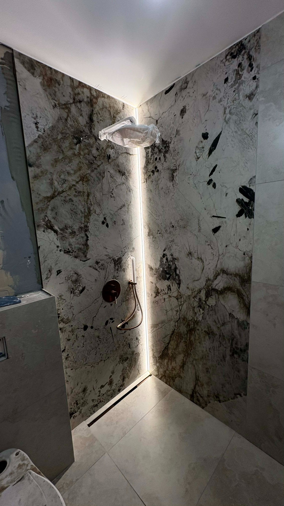 Wykończona kabina prysznicowa z marmurowymi płytami, podświetleniem LED i odpływem liniowym. Armatura w kolorze miedzi. Częściowo widoczna toaleta w folii ochronnej.