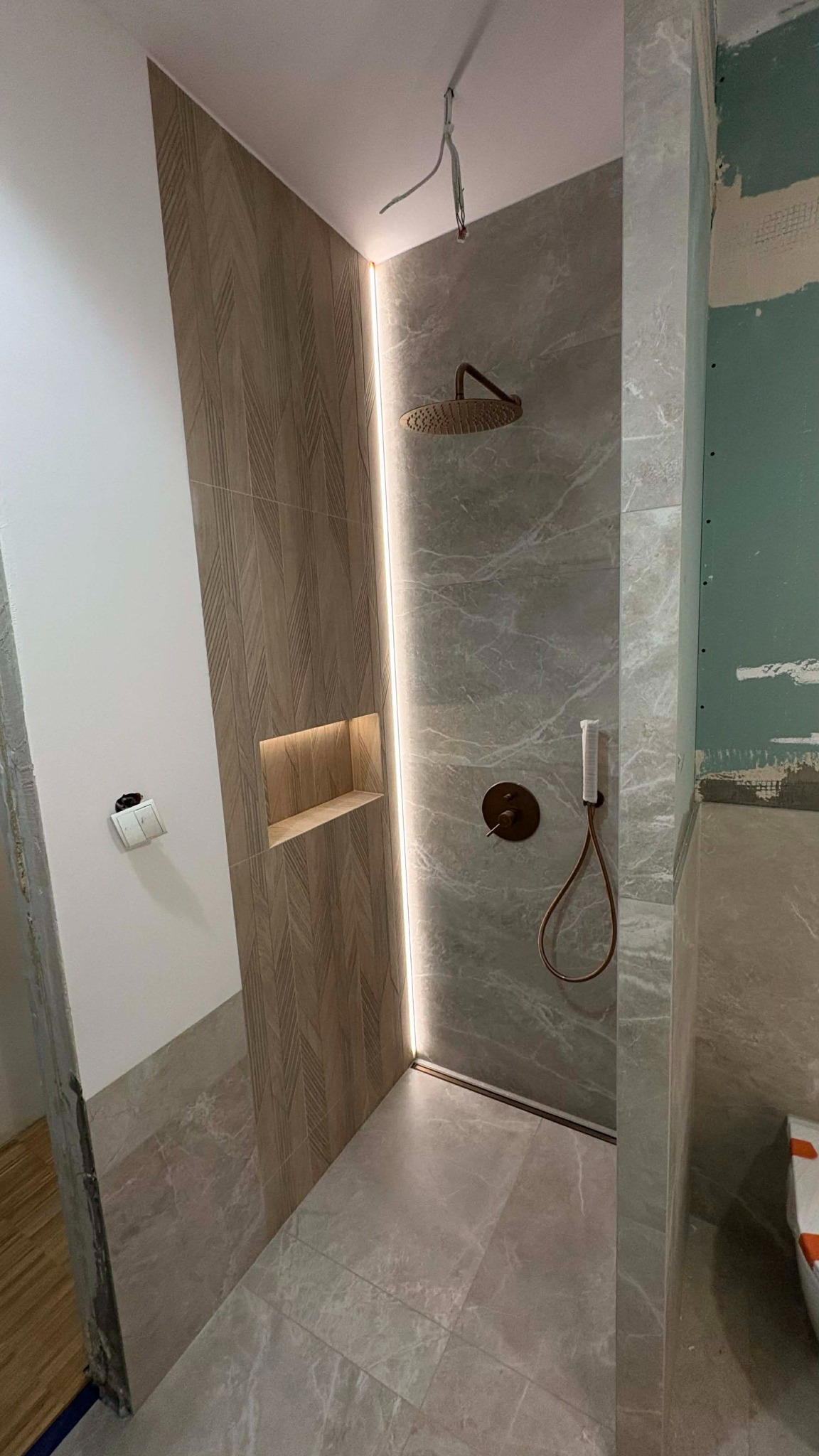 Wykończona kabina prysznicowa z oświetleniem LED i półką w Luboniu. Płytki imitujące drewno i marmur. Widoczne elementy instalacji wodnej w kolorze miedzi.