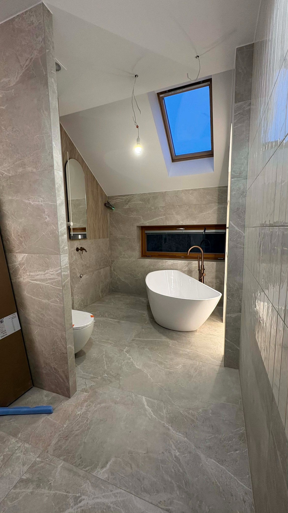 Nowoczesna łazienka na poddaszu z marmurowymi płytkami, wolnostojącą wanną i oknem dachowym. Widoczne niedokończone oświetlenie.
