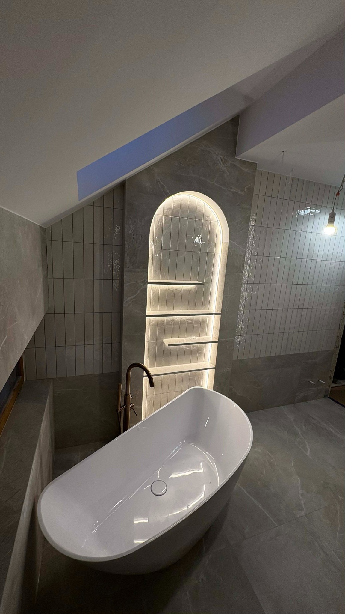 Nowoczesna łazienka w szarościach z wolnostojącą wanną i podświetlaną wnęką z półkami. Ukośny sufit dodaje unikalnego charakteru wnętrzu.