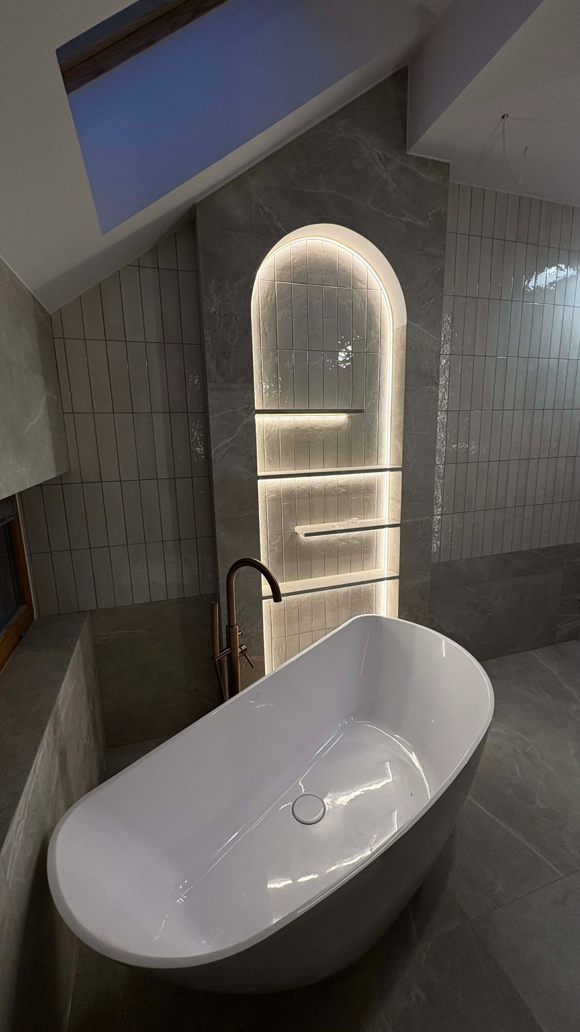 Nowoczesna łazienka na poddaszu z wolnostojącą wanną, złotą armaturą i podświetlaną wnęką z półkami wyłożoną pionowymi płytkami. Szare płytki na ścianach i podłodze.