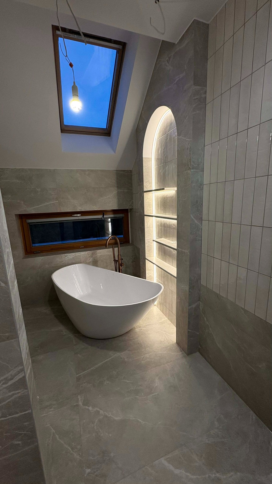 Nowoczesna łazienka z wolnostojącą wanną, oknem dachowym i podświetlaną wnęką. Płytki imitujące marmur na podłodze i ścianach. Bateria w kolorze miedzi.