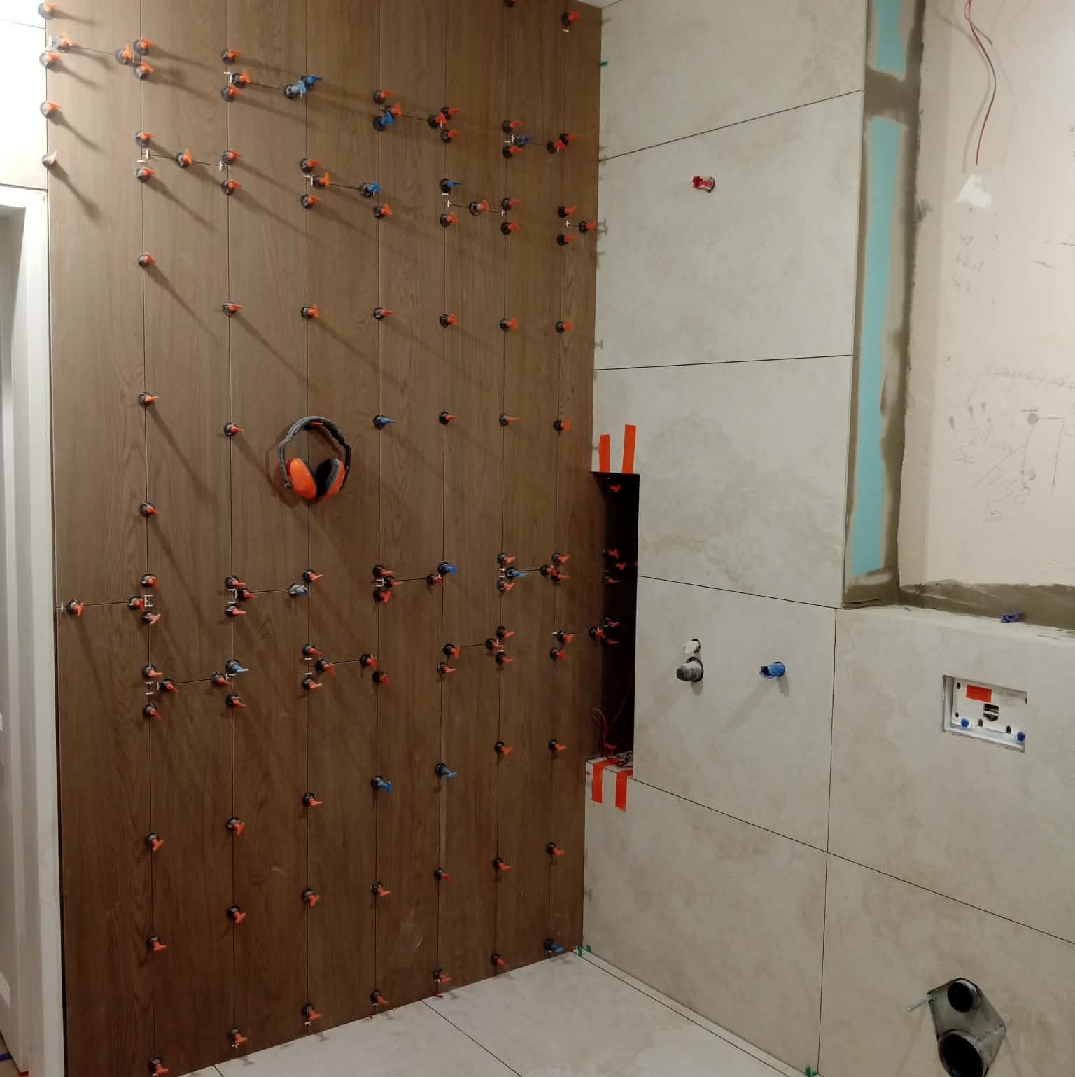 Układanie płytek imitujących drewno na ścianie z użyciem systemu poziomowania. Widoczne słuchawki robocze. Fragment ściany z jasnymi płytkami i instalacją.