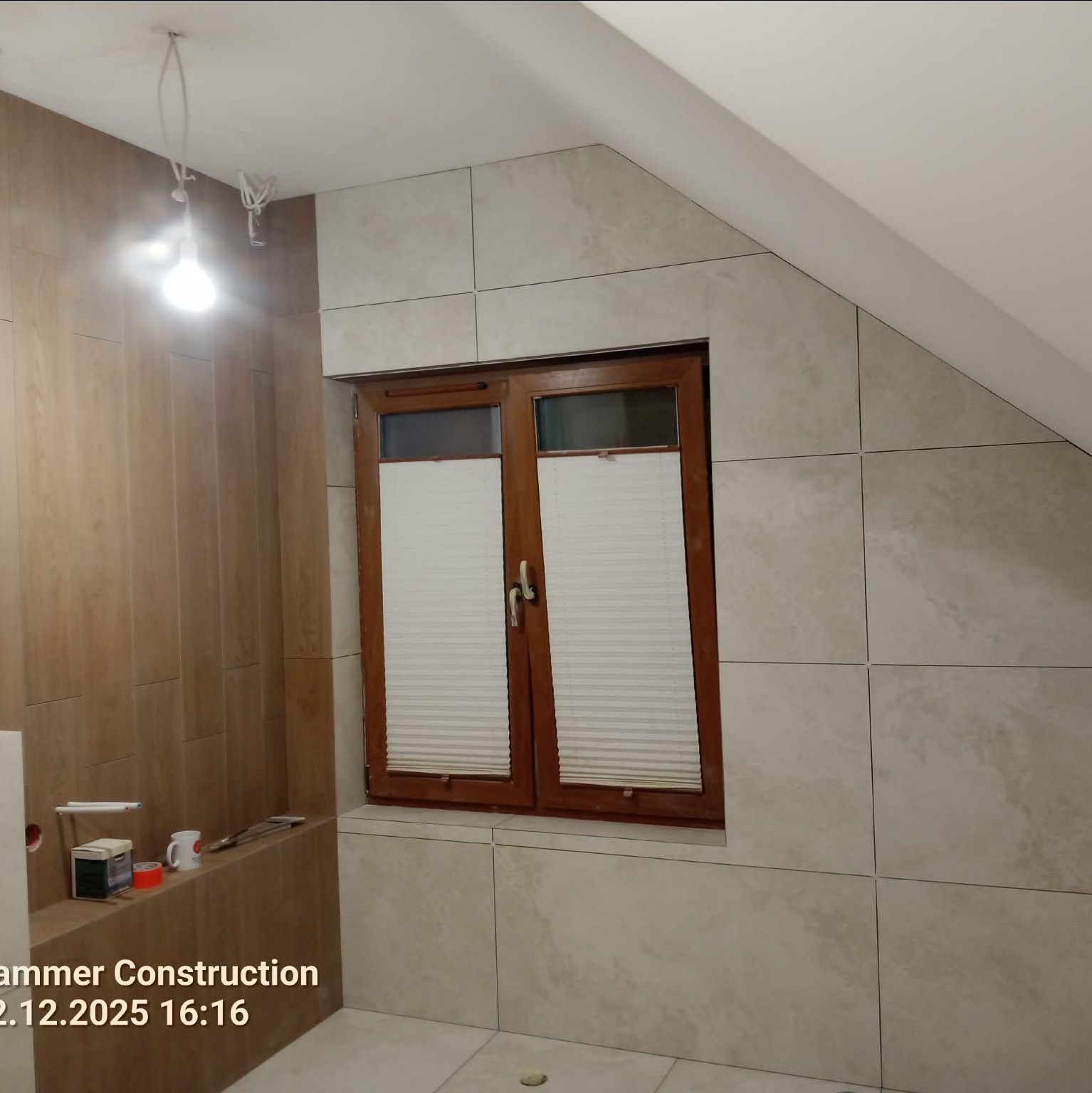 Wykończona łazienka na poddaszu z oknem w drewnianej ramie i beżowymi płytkami. Widoczne akcesoria łazienkowe i fragment instalacji. Ukończony etap prac glazurniczych.