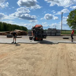 Układanie szarej kostki brukowej na piaskowym podłożu przy użyciu specjalistycznej maszyny; w tle stosy kostki, drzewa i błękitne niebo z chmurami.