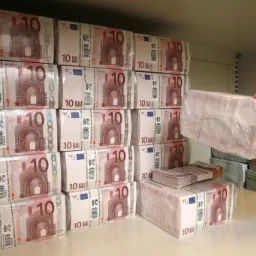 Rządki zapakowanych pakietów banknotów 10 euro na półce, z ręką sięgającą po kolejny pakiet, sugerujące dostępność gotówki.