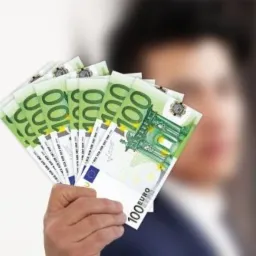 Mężczyzna w tle trzyma wachlarz banknotów 100 euro, lekko rozmazane tło.