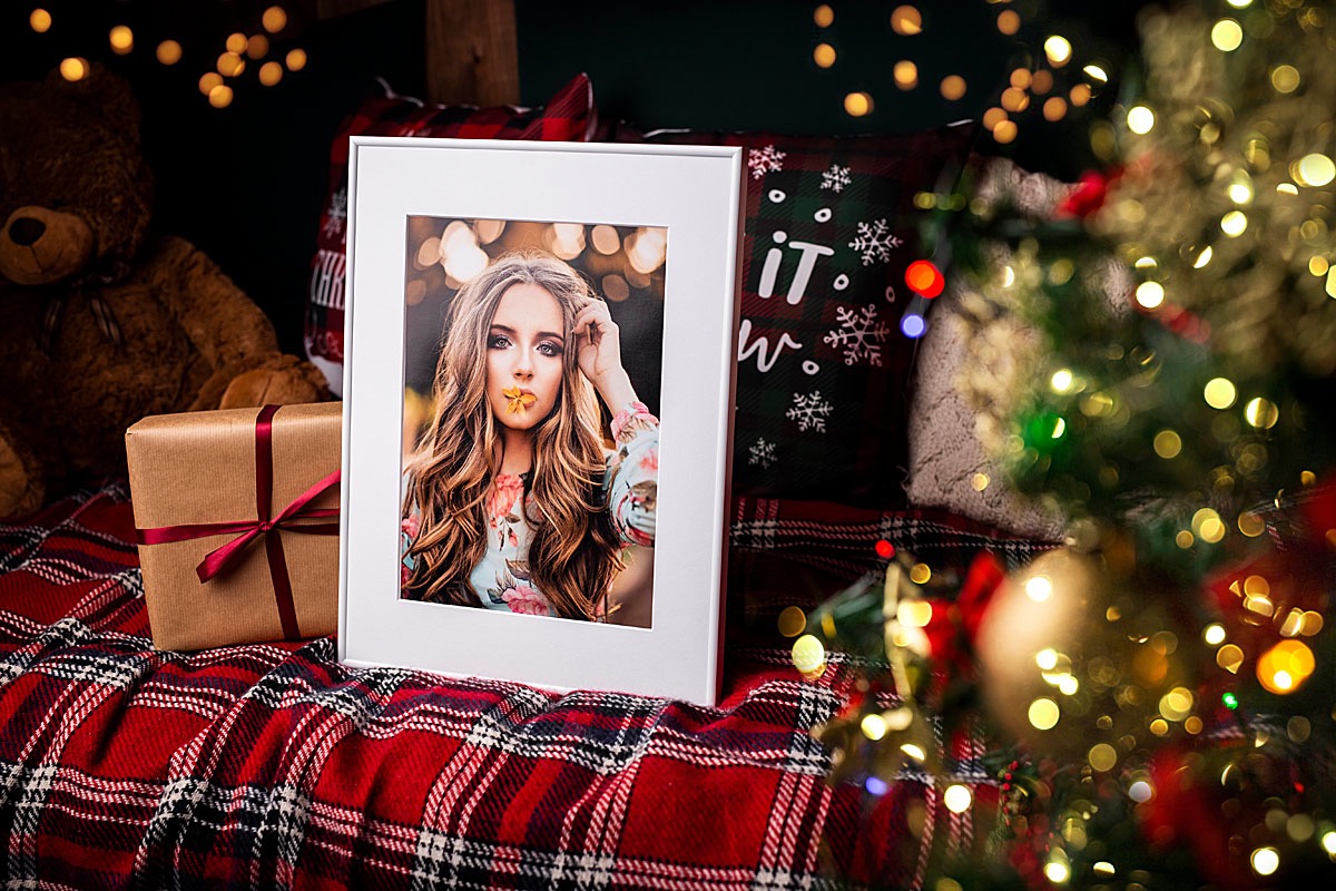 Oprawione portretowe zdjęcie młodej kobiety z kwiatem w ustach, leżące na czerwonej kraciastej tkaninie obok prezentu i pluszowego misia w świątecznej scenerii z choinką i lampkami.