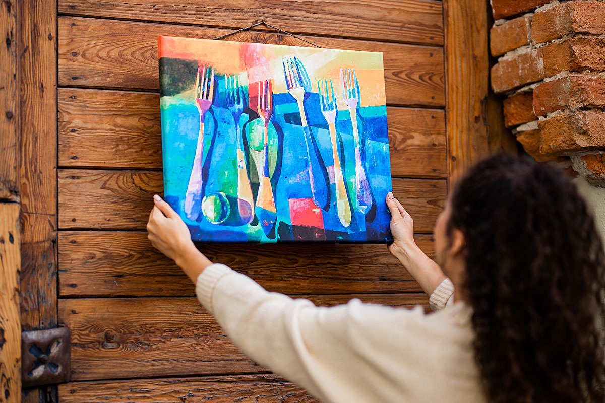 Kobieta zawiesza na drewnianej ścianie abstrakcyjny obraz przedstawiający kolorowe widelce na błękitnym tle.
